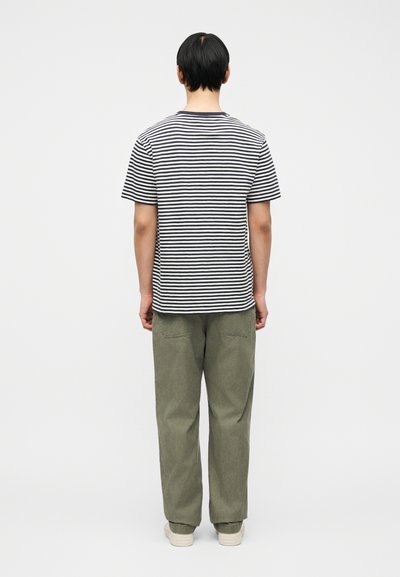 Άνδρας που στέκεται με την πλάτη, φοράει κοντομάνικο πουκάμισο με ασπρόμαυρες ρίγες, χακί παντελόνι και λευκά αθλητικά παπούτσια σε απλό φόντο.