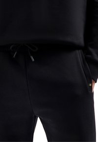 Schwarze Jogginghose aus weichem Stoff, mit elastischem Bund und Kordelzug, seitlichen Taschen und einem kleinen Logo-Detail auf einer Seite.