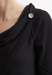 Svart stickad tröja med draperad halsringning, med en framträdande svart knapp på axeln. Mjuk textur och avslappnad passform.