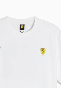 T-shirt blanc à col rond avec un petit logo jaune écusson Ferrari sur la poitrine et un logo Puma noir sur l'étiquette du col.