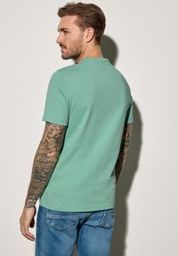 Mátově zelené bavlněné tričko s krátkým rukávem a kulatým výstřihem, nošeno s modrými džíny, na kterých jsou vidět propracované tetování na rukou.