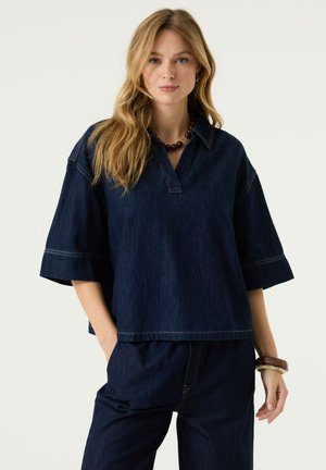Vrouw met lang blond haar draagt een losse donker denim top en broek, een bruine kralenketting en armbanden, staand tegen een effen achtergrond.