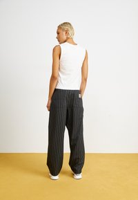 Mustat pinstripe-housut rentoa leikkuuta, joissa on kaksi takataskua ja pieni brändilappu. Yhdistetty valkoiseen ilman hihoja olevaan paitaan.
