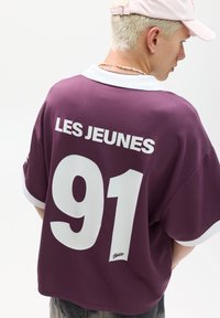 Burgundy oversized t-shirt featuring white collar and large "LES JEUNES" text and "91" number on the back, smooth fabric with short sleeves.