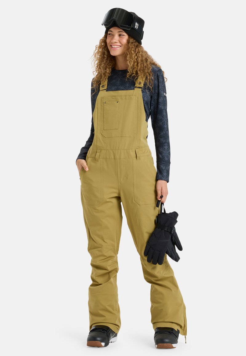 Žlté snehové nohavice na traky s predným vreckom a širokými nohavicami. Kombinované s čiernymi rukavicami a vzorovanou košeľou s dlhým rukávom.