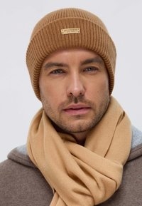 Brown ribbed knit beanie with a metal logo tag, paired with a light brown scarf, worn over a gray hooded top. Smooth texture throughout.