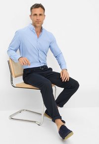 Lichtblauw overhemdd blouse, marineblauwe broek. Soepele stof met een subtiele textuur. Zittend op een metalen stoel met een houten zitting. Donker schoeisel.