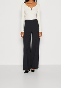 Donna che indossa un top bianco a maniche lunghe con zip frontale e pantaloni neri a vita alta e gamba larga, in piedi contro uno sfondo bianco.