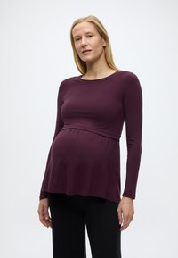 Έγκυος γυναίκα με μακριά ξανθά μαλλιά, φορώντας μωβ μπλούζα με μακριά μανίκια και μαύρο παντελόνι, στέκεται μπροστά σε ένα απλό λευκό φόντο.