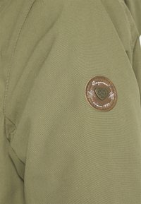 Tela de chaqueta verde oliva con textura suave; presenta un parche circular marrón con un diseño de corazón y el texto "Raquetas desde 1997."
