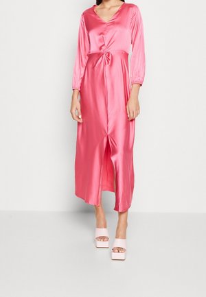 Kobieta ubrana w błyszczącą różową sukienkę midi z satyny z długimi rękawami, marszczoną talią, przednim rozcięciem i różowymi sandałami na platformie z odkrytymi palcami.