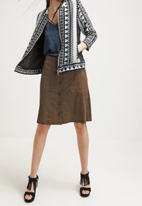 Zwart-wit gestreepte jas, marineblauwe top, bruine suède rok met knopen aan de voorkant, zwarte fringed haksandalen. Eenvoudige achtergrond.