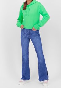 Persona che indossa un vivace felpa verde, jeans a campana blu con fessure laterali e sneaker bianche con dettagli rosa, in piedi con le mani nelle tasche.