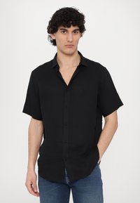 Μαύρο κοντομάνικο πουκάμισο με κουμπιά σε ελαφρύ ύφασμα, με κλασικό γιακά και χαλαρή εφαρμογή. Φορεμένο με μπλε τζιν.