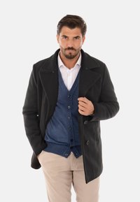 Cappotto in misto lana nero con bottoni doppiopetto, indossato sopra un cardigan blu, una camicia bianca e pantaloni beige. Tessuto liscio e taglio su misura.