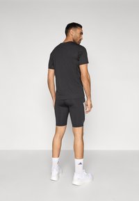 Schwarzes Sport-T-Shirt und enganliegende Shorts, beide in dunklen Tönen mit weißen Seitenstreifen. Weiße Sneaker und Socken komplettieren das Outfit.