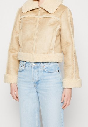 Femme portant une veste en peau retournée beige avec une fermeture éclair à l'avant et un jean taille haute bleu clair, debout devant un fond blanc.