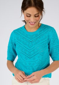 Suéter de punto en color verde azulado con mangas cortas, que presenta un patrón geométrico de calado y un cuello y puños acanalados, combinado con pantalones claros.