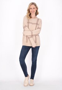 Beige Strickpullover mit braunem Karomuster, langen Ärmeln, geripptem Ausschnitt, kombiniert mit dunkelblauen Skinny-Jeans und hellen Sneakers.