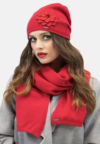 Czerwony, dziergany beanie z filcowym akcentem w postaci kwiatka oraz dopasowanym czerwonym szalikiem. Materiał jest miękki, a kształty są zorganizowane i przytulne.
