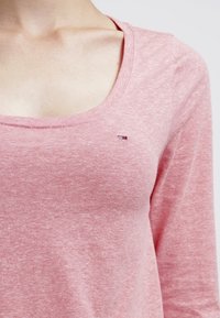 Ljusrosa långärmad topp med rundad halsringning, tillverkad av mjukt tyg. Har en liten logotypdekoration nära halsringningen.