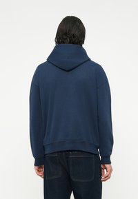 Mornarsko moder hoodie izdelan iz mehkega materiala, z prostornim kapuco, zasnovo z nizkimi rameni in rebrastimi robovi na dnu ter rokavih.