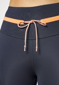 Marinblå leggings med en orange dragsnodd. Midjebandet har en slät yta med ett subtilt marmorerat mönster. Förstärkta sömmar finns.
