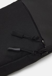 Svart stoffpose med teksturert overflate, doble glidelåser med dragremmer, og en skrå design på den ene siden.