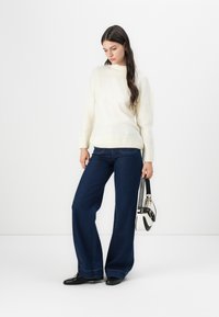 Off-white gestrickter Pullover mit geripptem Saum, getragen mit dunkelblauen weiten Jeans. Schwarze Schuhe und eine schwarz-weiße Handtasche sind dabei.