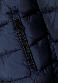 Navy blue puffer jacket with a zippered pocket, featuring quilted sections and a smooth texture. Zipper has a black pull tab.