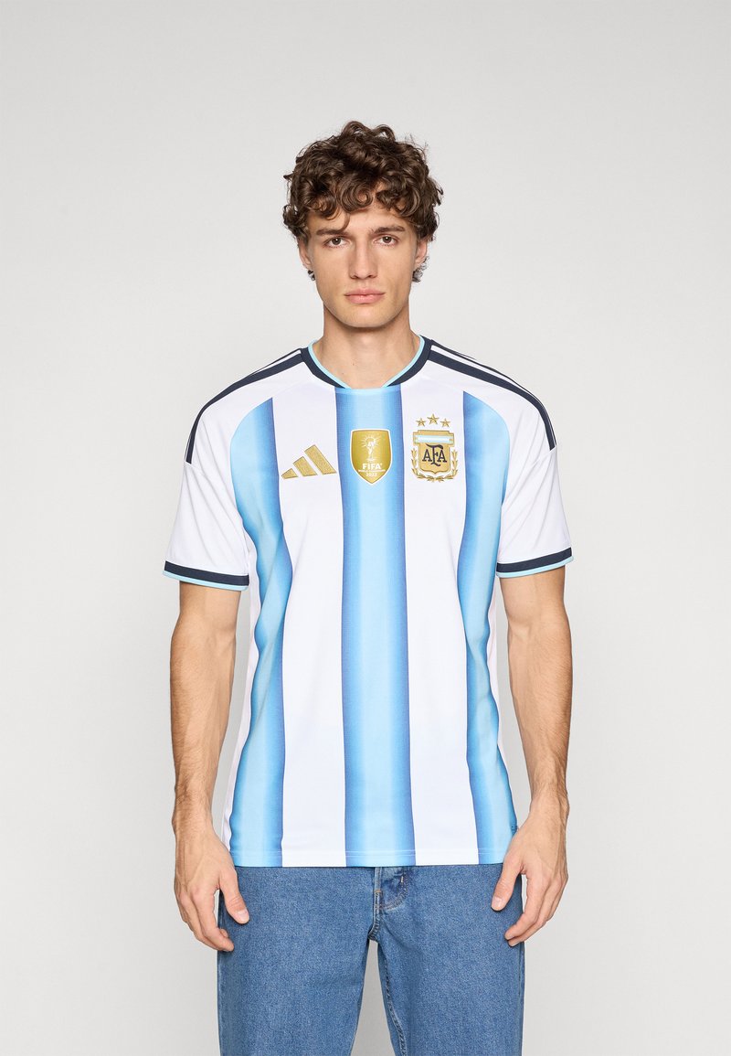 Man som bär Argentinas fotbollslandslagströja med blå och vita ränder, FIFA 2022-märke och Adidas-logotyp, tillsammans med blå jeans.