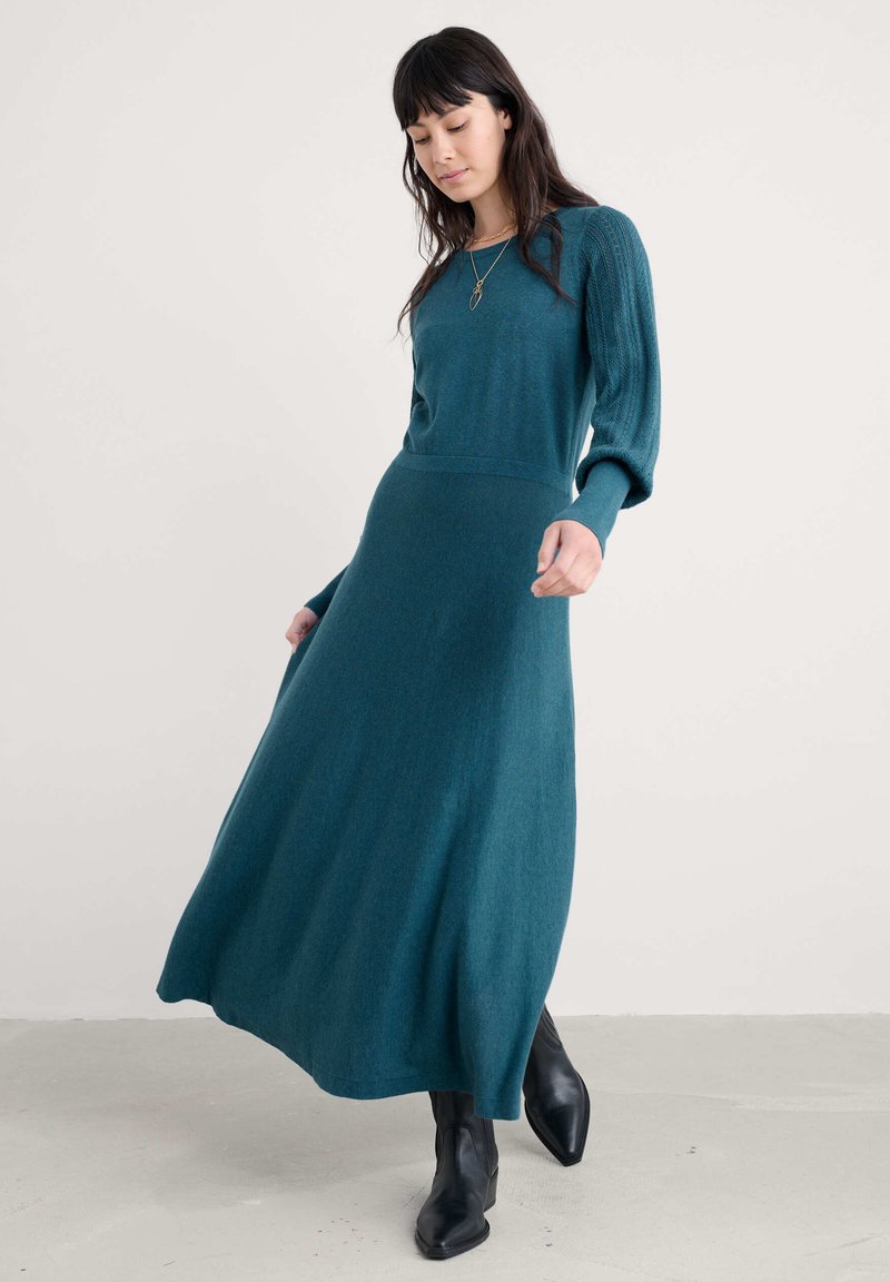 Mėlynos žalios spalvos megztas suknelė su ilgomis rankovėmis, raukšlėtomis detalėmis ir laisvai krentančia sijonine dalimi. Derinama su juodomis aulinėmis batais. Fainos tekstūros, kasdieniškas siluetas.