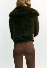 Donna che indossa un giubbotto di pelliccia marrone scuro e peloso, pantaloni marroni testurizzati e occhiali da sole tartarugati, vista di spalle su uno sfondo bianco.