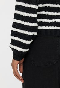 Hånd hvilende nær hoften, iført svart-hvitt stripet strikkegenser og svarte bukser med synlig baklomme.