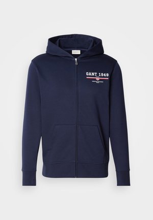 Sweat à zip bleu marine en tissu doux, avec le logo "GANT 1949" en blanc et rouge. Comprend des poches avant et une capuche avec cordon de serrage.
