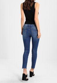 Pantalones vaqueros ajustados de color azul con una textura desgastada, diseño de cinco bolsillos y ligeros desgarros en los dobladillos. Combinados con sandalias de tacón alto negras.