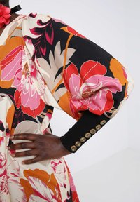 Floral jurk met levendige rode, roze en oranje bloemen op een crèmekleurige achtergrond, met zwarte mouwen en knopenaccenten op de manchetten.
