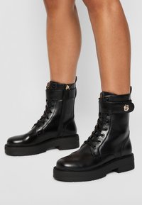 Botas de tobillo de cuero negro con cordones, suela gruesa, cremallera lateral y una correa con un adornos metálico decorativo. Textura suave y diseño estilizado.