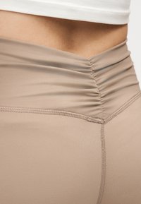 Beige Leggings mit hohem Bund und gerafftem Detail am hinteren Bund, getragen mit einem kurzen weißen Top, das den Bauch freilegt.