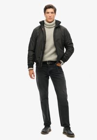 Chaqueta bomber negra sobre un suéter de cuello alto gris claro, combinada con jeans oscuros y zapatos negros. La chaqueta cuenta con puños y cuello acanalados.
