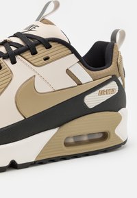 Baskets Nike Air Max beige et noires avec coussin d’air visible, lacets noirs et languettes à tirer au talon et sur la languette.