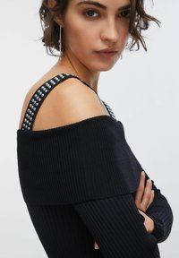 Črno rebrasto top brez naramnic z vzorčastimi trakovi, ki imajo srebrne poudarke. Tkanina ima teksturirano površino, ki ustvarja oprijeten oprijem.