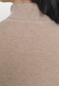 Pull beige en tricot à col roulé avec un col côtelé et une texture douce, une finition lisse et des variations subtiles de couleur.