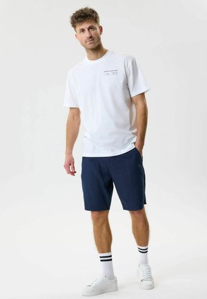 Bărbat stând într-un tricou sport alb, pantaloni scurți bleumarin, adidași albi și șosete albe cu dungi negre pe un fundal simplu.