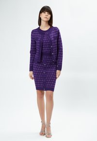 Conjunto de cárdigan y falda de punto morado con un patrón geométrico, con detalles de botones y dobladillos acanalados. Sandalias de tiras completan el look.
