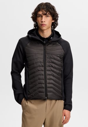 Ung mand med krøllet hår iført en sort lynlåset hættetrøje og beige bukser, stående mod en ensfarvet lys baggrund.