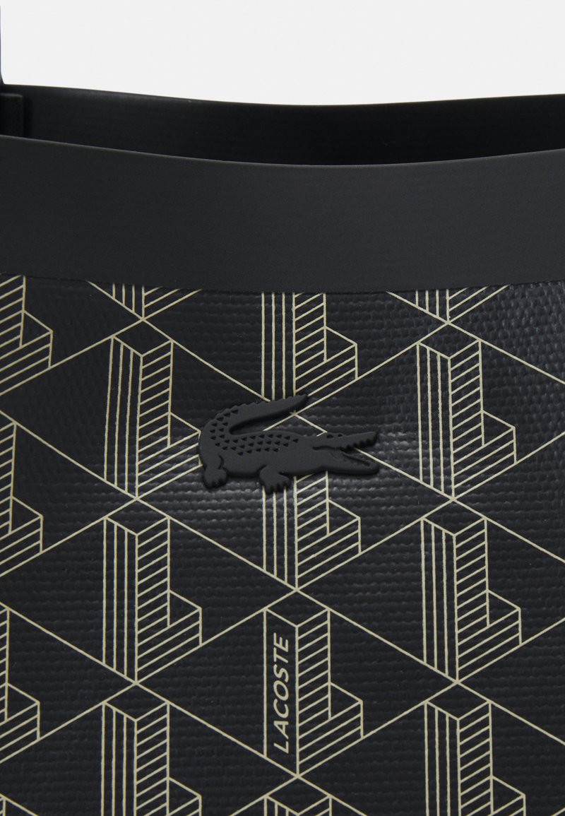 Czarna torba o teksturowanej powierzchni z geometrycznym złotym wzorem, zawierająca czarne logo krokodyla oraz słowo "LACOSTE" wkomponowane w design.