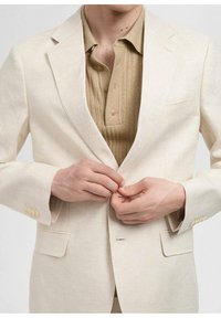 Blazer din in bej deschis, cu rever tăiat, două buzunare frontale și închidere cu nasture. Sub acesta, se vede un tricou polo cu nervuri de culoare bej.