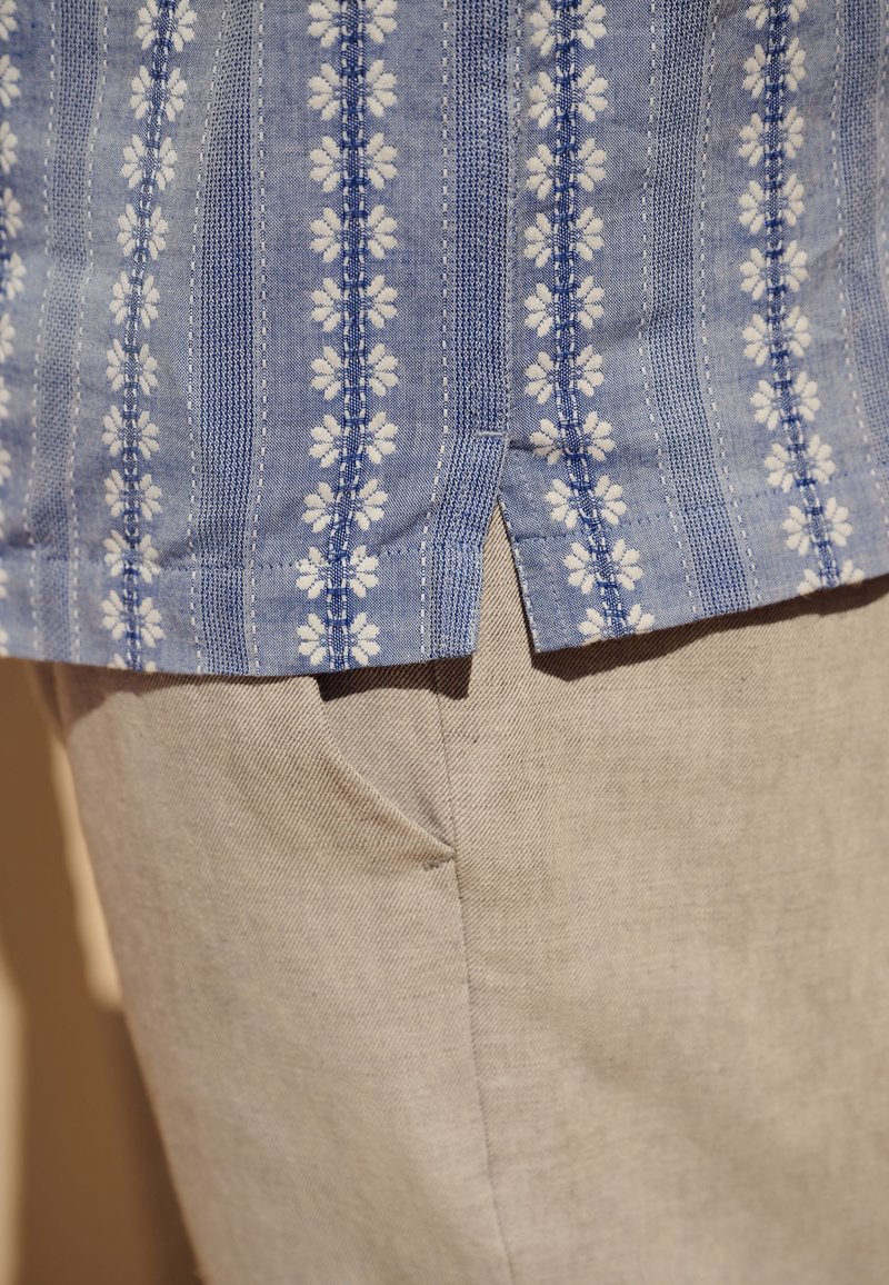 Chemise bleue à motif avec des rayures florales blanches portée sur un pantalon beige, montrant une couture latérale et une petite fente à l'ourlet de la chemise.