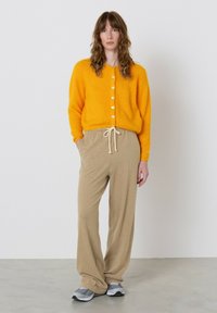 Geltonas pūkuotas kardiganas su baltais mygtukais, derinamas su kreminės spalvos kelnėmis su raišteliais ir pilkomis sportinėmis avalynėmis, ant paprasto fono.